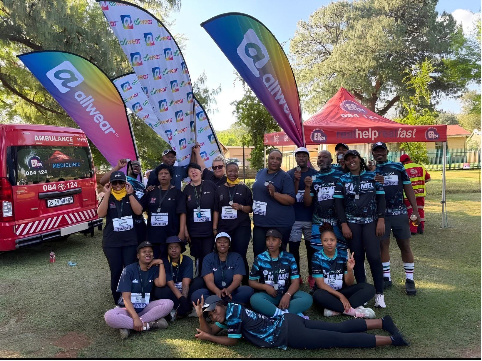 A relay team in matching shirts gathers beside the ambulance and sponsor flags at the Memel Walk